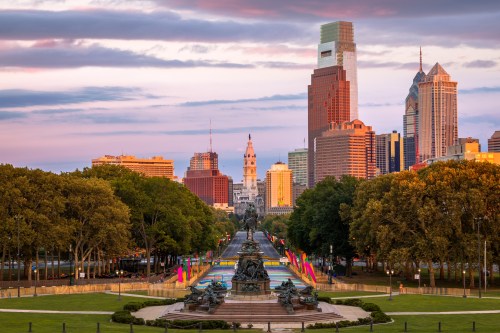 A vibrant cityscape of Philadelphia at sunset, featuring skyscrapers and lush trees along a scenic parkway.