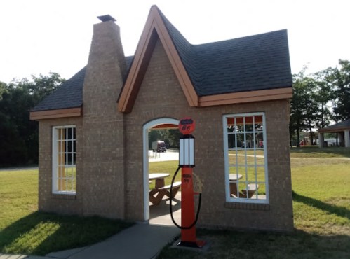 A small brick building with a peaked roof and a vintage gas pump outside, set in a grassy area.