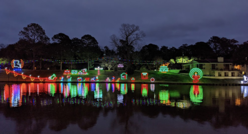 Colorful holiday lights reflect on a calm lake, illuminating trees and festive decorations along the shore at night.
