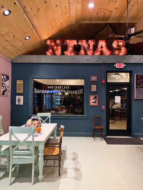 A cozy interior of a restaurant with a large "NINAS" sign, wooden ceiling, and colorful chairs around a table.