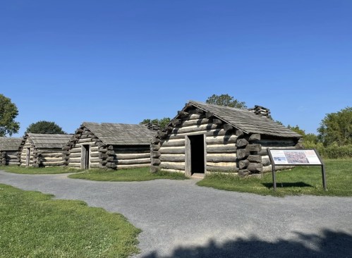 Log cabins with slanted roofs along a gravel path, set against a clear blue sky and green grass.