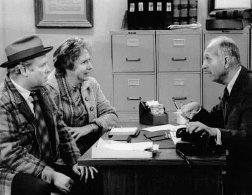 A black-and-white scene of three people at a desk, engaged in a serious conversation. Two are seated, one is standing.