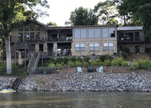 A large stone house with multiple windows overlooks a lake, surrounded by greenery and a rocky shoreline.