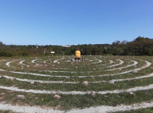 A person sits on a bench in the center of a circular stone labyrinth, surrounded by greenery and a clear blue sky.