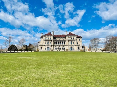 A grand mansion with a red-tiled roof, set against a blue sky with clouds, surrounded by a lush green lawn.