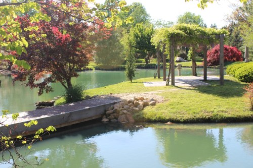 A serene pond surrounded by lush greenery and colorful trees, featuring a wooden dock and a pergola on the shore.