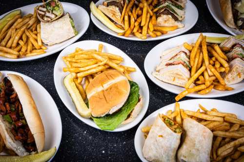 A variety of sandwiches and wraps served with golden fries on white plates, with pickles on the side.