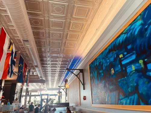 Interior of a restaurant featuring a decorative ceiling, colorful flags, and a large blue painting on the wall.