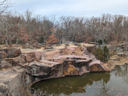 Rocky landscape with a calm pond surrounded by bare trees and autumn foliage under a cloudy sky.