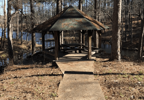 A wooden gazebo in a wooded area by a pond, with a path leading up to it and trees surrounding the scene.