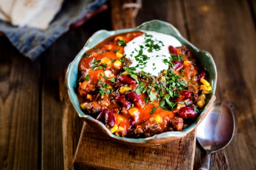 A bowl of chili topped with sour cream and fresh herbs, served on a wooden surface with a spoon nearby.