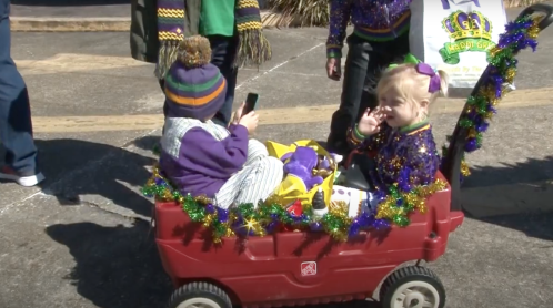 Two children sit in a decorated red wagon, one using a phone, surrounded by festive Mardi Gras decorations.