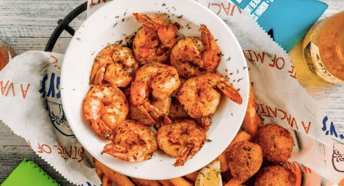 A bowl of seasoned shrimp served with fries and hushpuppies, accompanied by a drink in the background.