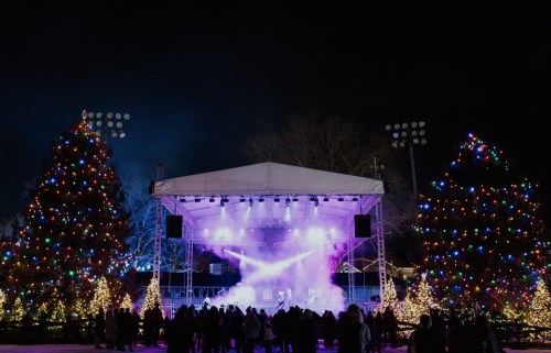 A festive outdoor stage with colorful lights, surrounded by decorated trees and an audience enjoying a nighttime event.