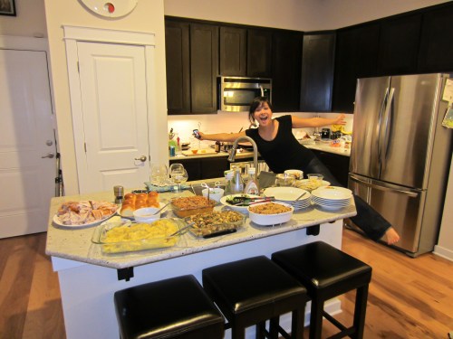A woman playfully poses next to a kitchen island filled with various dishes and a festive spread in a modern kitchen.