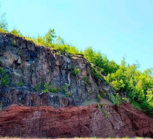 A rocky cliff with layered red and gray stone, topped with lush green trees under a clear blue sky.