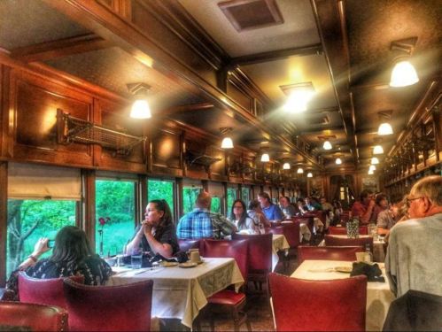 A vintage dining car filled with diners enjoying meals, surrounded by wooden decor and large windows with greenery outside.