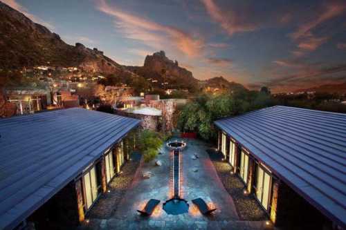 A serene outdoor space with modern buildings, a central water feature, and mountains under a colorful sunset sky.