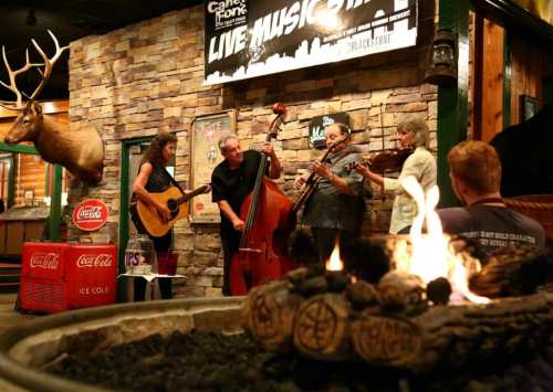 A live music performance in a rustic venue, featuring musicians with a guitar, bass, and violins, with a fire pit in the foreground.