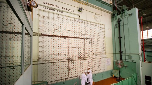 Two workers in white uniforms stand in front of a large control panel in a graphite reactor facility.