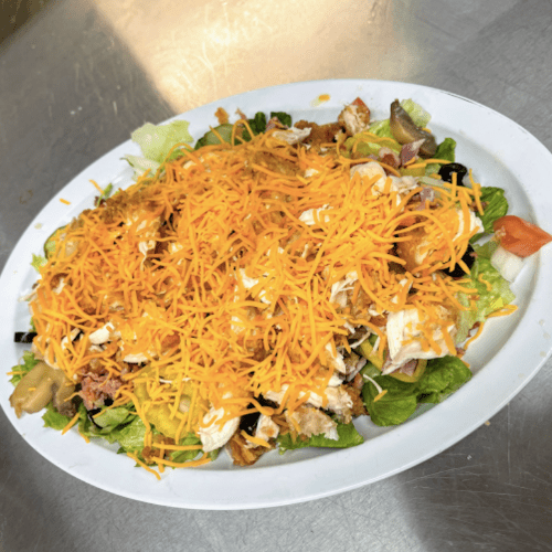 A large salad topped with shredded cheese, lettuce, tomatoes, and various toppings on a white plate.