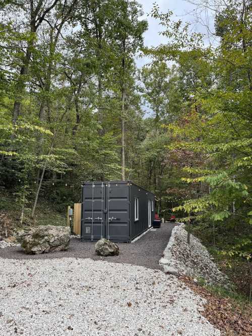 A modern black container home nestled in a wooded area, surrounded by gravel and greenery.