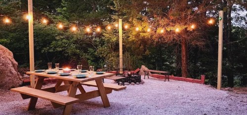 A cozy outdoor dining area with a wooden picnic table, string lights, and a fire pit surrounded by trees.