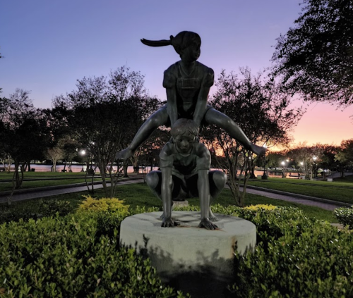 A bronze sculpture of a girl on a boy's back, set in a park during sunset with trees and lights in the background.
