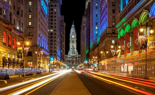 A vibrant city street at night, featuring illuminated buildings and a clock tower in the distance, with light trails from traffic.