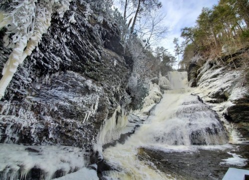 A frozen waterfall cascading down rocky cliffs, surrounded by icy formations and trees in a winter landscape.