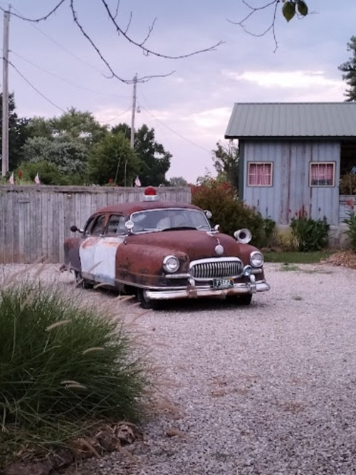 A vintage, rusty police car parked on gravel, with a small shed and greenery in the background.