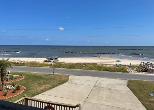 A scenic view of a beach with calm waters, a sandy shore, and a clear blue sky, seen from a nearby house.