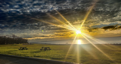Sunrise over a calm lake, with picnic tables in the foreground and dramatic clouds in the sky.