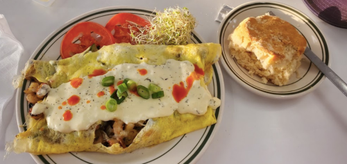 A plate featuring a large omelet with toppings, sliced tomatoes, sprouts, and a biscuit on the side.
