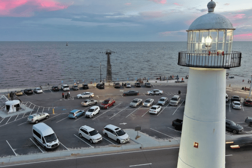 A lighthouse stands by the shore, overlooking a parking lot filled with cars and a calm sea under a colorful sky.