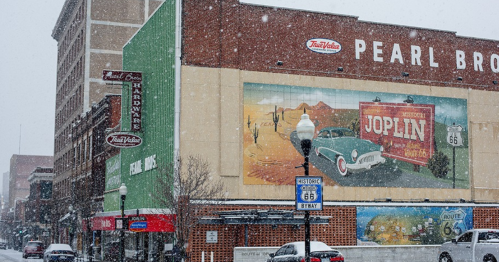 A snowy street scene featuring a historic building with a colorful mural and a Route 66 sign in Joplin, Missouri.