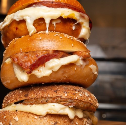 Three stacked bagels filled with melted cheese, bacon, and a sweet potato topping, drizzled with sauce.