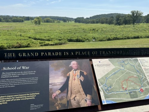 A scenic view of a battlefield with a sign about the Grand Parade and a portrait of a historical figure.