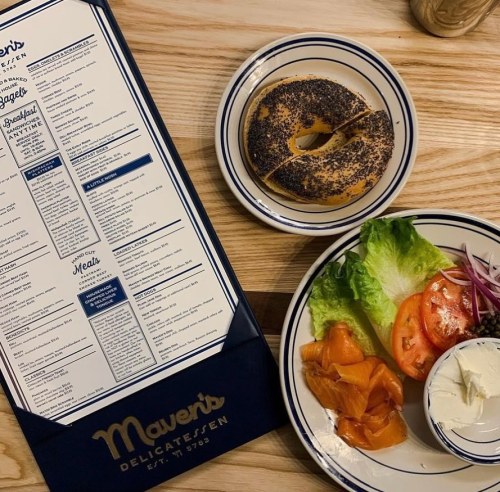 A menu beside a plate with a bagel, smoked salmon, lettuce, tomato, onion, and a small bowl of cream cheese.