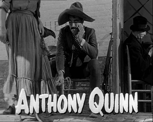 A black-and-white scene featuring a man in a sombrero and suit, sitting with a woman in a long dress, with the name "Anthony Quinn" displayed.