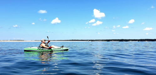 A person relaxes in a kayak on calm water, surrounded by blue skies and fluffy clouds.