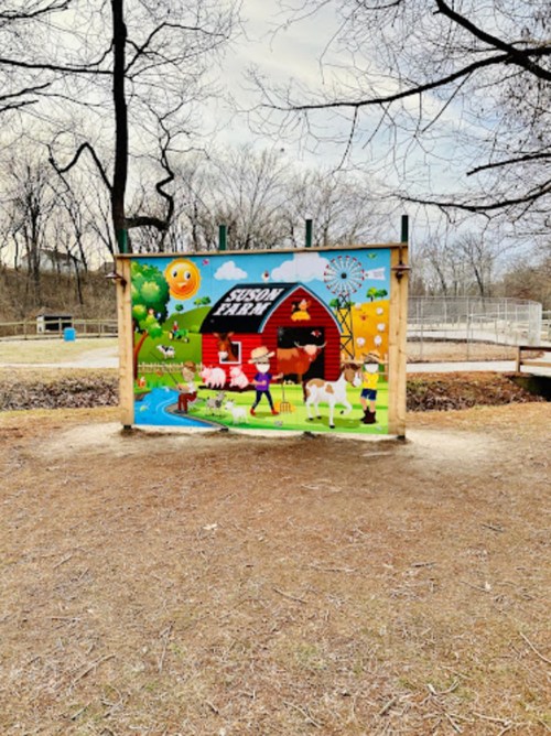 Colorful mural of a farm scene with a barn, animals, and cartoon characters, set in a park with trees in the background.