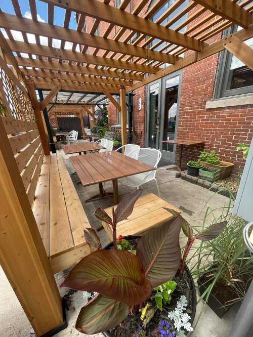 Outdoor seating area with wooden pergola, tables, and potted plants against a brick wall. Bright and inviting atmosphere.