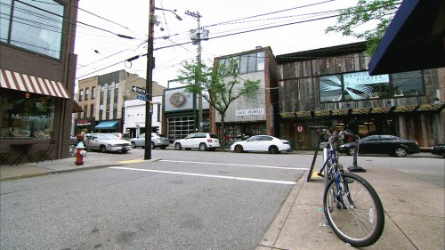 A street scene featuring shops, parked cars, and a bicycle leaning against a pole on a sunny day.