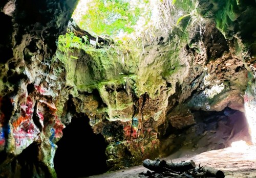 A rocky cave interior with green moss, sunlight streaming in from above, and colorful graffiti on the walls.