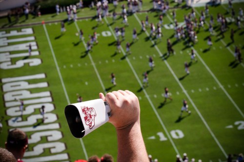 A hand holds a megaphone above a football field with players warming up in the background.