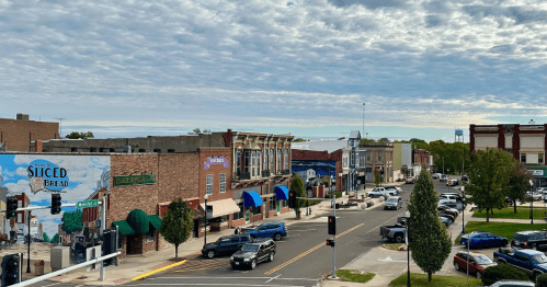 A view of a small town street with shops, murals, and cloudy skies, featuring parked cars and traffic lights.