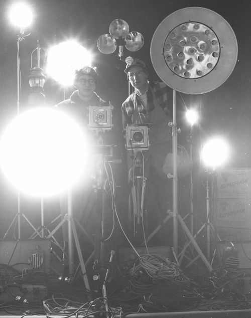 Two photographers stand behind large studio lights and cameras, surrounded by bright lighting equipment.