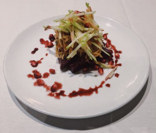 A gourmet dish featuring layered vegetables, topped with green apple strips and pomegranate seeds on a white plate.