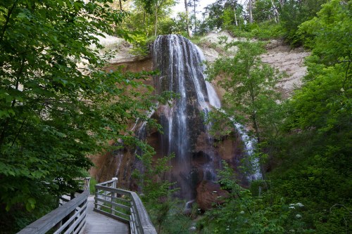 A serene waterfall cascading down a rocky cliff, surrounded by lush green trees and a wooden walkway.